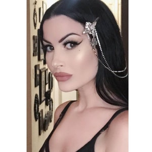 Puede incluir: Una pinza para el cabello en tono plateado con un dije central en forma de cruz adornado con pedrería transparente. El accesorio incluye dos delicadas cadenas plateadas colgantes decoradas con pequeños brillantes que caen por el cabello.