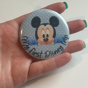 May include: A round button with a light blue background and white polka dots. The button features a cartoon image of Mickey Mouse and the text "My first Disney trip."