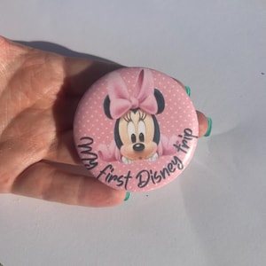 May include: A pink button with a Minnie Mouse graphic. The button features Minnie Mouse with a pink bow and the text "My first Disney trip" in a script font. The background is a light pink with white polka dots.