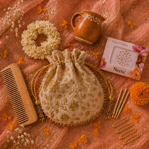 May include: An assortment of Indian wedding accessories on a peach-colored fabric. Items include a floral hairpiece, a small ceramic mug, a drawstring pouch with gold floral embroidery, a wooden comb, hairpins, and a box of face jewels.