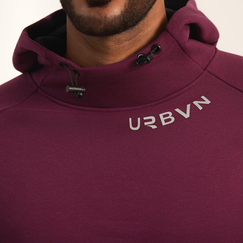 May include: A close-up of a maroon hoodie with a high collar and a drawstring. The word "URBVN" is printed in silver on the front. The hoodie has a black lining in the hood and a modern, athletic design.