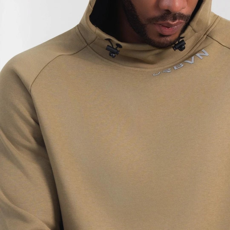 May include: A tan hoodie with a high collar and a black hood lining. The collar features black hardware and the word "URBBN" in silver lettering. The hoodie is made of a soft, comfortable material.