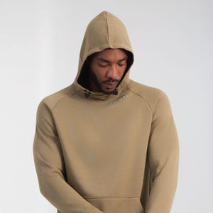 May include: A tan hoodie with a drawstring hood and a kangaroo pocket. The hoodie has a high neckline and the word "NAVN" is printed on the chest. The sleeves are long and the material appears to be a soft knit.