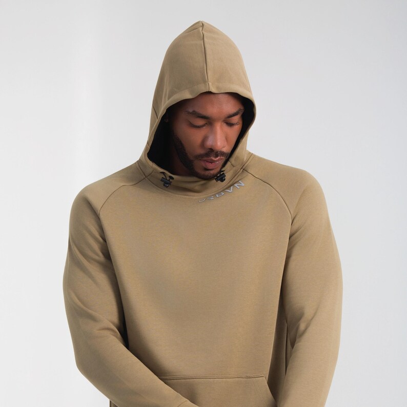 May include: A tan hoodie with a drawstring hood and a kangaroo pocket. The hoodie has a high neckline and the word "NAVN" is printed on the chest. The sleeves are long and the material appears to be a soft knit.