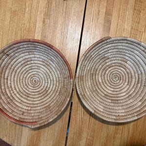 May include: Two round, shallow woven baskets featuring a coiled construction pattern. The baskets are crafted from natural light-colored fibers with subtle variations in tone, including sections of reddish-brown accents along the outer rims.
