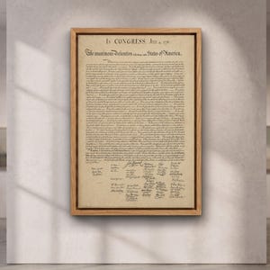 Könnte beinhalten: Eine gerahmte Nachbildung der Unabhängigkeitserklärung auf gealtertem, pergamentartigem Papier mit schwarzem Drucktext und Unterschriften. Das Dokument trägt den Titel In Congress, July 4, 1776, und ist in einem hellen Holzrahmen gefasst.