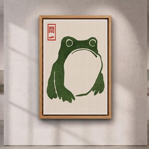 May include: A framed wall art print featuring a dark green frog illustration on a light cream background. In the upper left corner, there is a red square stamp containing the Japanese characters 問一. The artwork is encased in a light wood frame.
