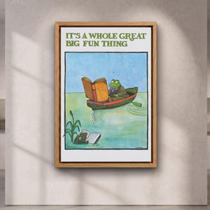 Peut inclure: Une impression d'art encadrée représentant une grenouille verte dans une petite barque lisant un grand livre, avec le texte IT'S A WHOLE GREAT BIG FUN THING en haut. Sous la barque, une autre grenouille lit un livre près de roseaux verts.
