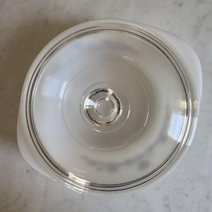 May include: A round white milk glass casserole dish with a clear glass lid. The lid features a central circular knob and a smooth rim. The base of the dish has integrated side handles, and the overall design is simple, functional, and clean.