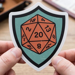Op de afbeelding: Een schildvormige sticker met een lichtblauwe achtergrond en een dikke zwarte rand. In het midden staat een afbeelding van een oranje twintigzijdige dobbelsteen met zwarte cijfers, waaronder een opvallende 20, 18, 13, 9, 16, 8, 12 en 2.