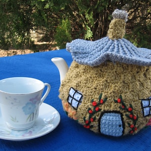 Puede incluir: Una funda de tetera de ganchillo con forma de casa con un techo azul, ventanas y una puerta. La funda está decorada con bayas rojas y flores. Una taza de té y un platillo blancos están en primer plano.
