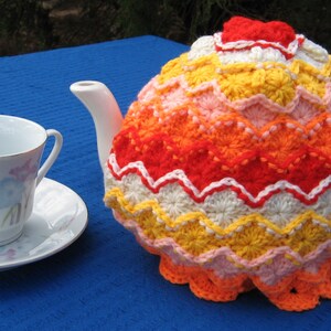 May include: A crocheted teapot cozy in shades of orange, yellow, pink, and white. The cozy features a scalloped edge and a repeating pattern of flowers.