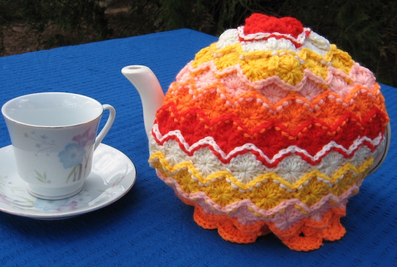 May include: A crocheted teapot cozy in shades of orange, yellow, pink, and white. The cozy features a scalloped edge and a repeating pattern of flowers.