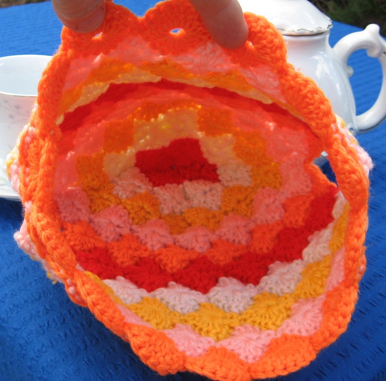 May include: A crocheted tea cozy in shades of orange, yellow, pink, and white. The cozy is made with a repeating pattern of small, rounded shapes.