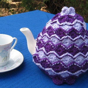 May include: A purple and white crocheted teapot cozy with a scalloped edge. The cozy is on a white teapot with a white teacup and saucer on a blue surface.