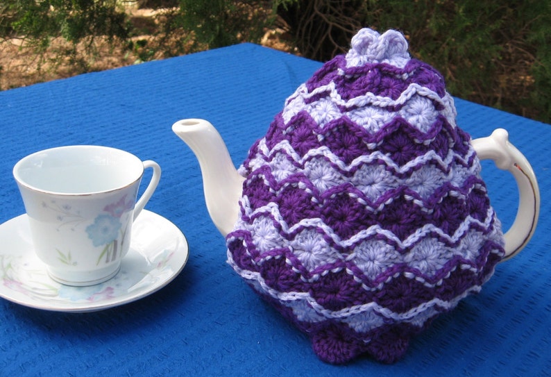 May include: A purple and white crocheted teapot cozy with a scalloped edge. The cozy is on a white teapot with a white teacup and saucer on a blue surface.