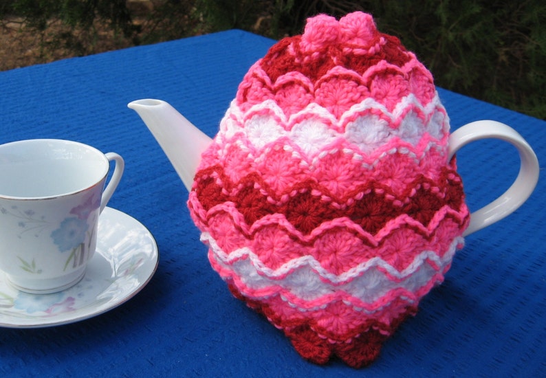 May include: A crocheted teapot cozy in pink, white, and red with a scalloped design. The cozy is on a blue surface.
