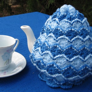 May include: A blue and white crocheted teapot cozy with a scalloped edge. The cozy is on a white teapot with a handle and spout.