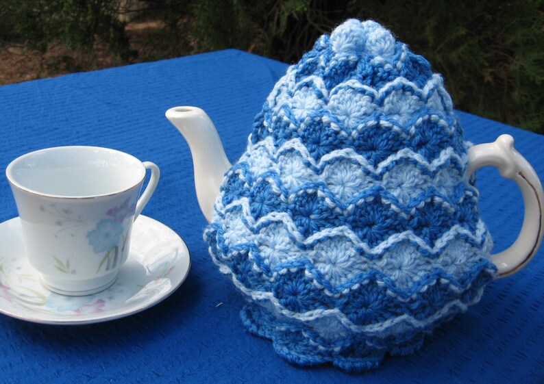 May include: A blue and white crocheted teapot cozy with a scalloped edge. The cozy is on a white teapot with a handle and spout.