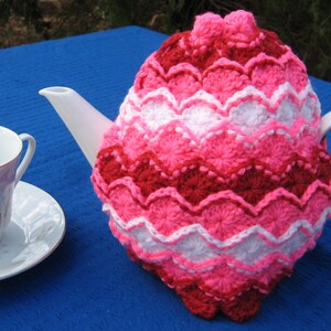 May include: A crocheted teapot cozy in pink, white, and red with a scalloped design. The cozy is on a blue surface.