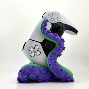 Könnte beinhalten: Ein weiß-schwarzer Videospiel-Controller mit einem lila-grünen Design, das von einem Oktopus inspiriert wurde. Der Controller hat einen schwarzen Joystick und Tasten mit einer lila Tentakelbasis.
