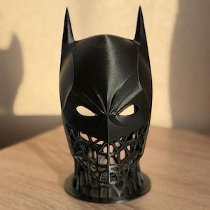 May include: A black Batman mask with pointed ears and a detailed, open-mouth design. The mask is 3D-printed and sits on a round base. The eye openings are visible.