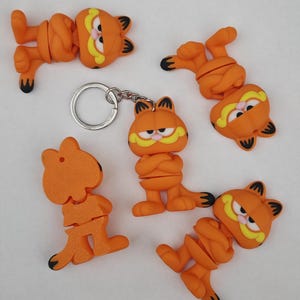 May include: Orange Garfield cat keyrings and charms. The keyrings and charms feature the cartoon cat with black stripes, yellow moustache, and a grumpy expression. The charms are in various poses, some with a keyring.