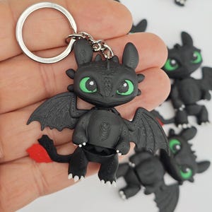 Könnte beinhalten: Schwarzer Drachen-Schlüsselanhänger mit grünen Augen und roten Schwanzakzenten. Der Drache hat detaillierte Flügel und ist an einem silbernen Schlüsselring befestigt. Mehrere ähnliche Schlüsselanhänger sind im Hintergrund zu sehen.