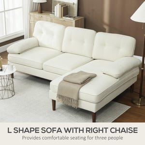 May include: Cream L-shaped sofa with a right chaise. The sofa features tufted back and seat cushions, and a matching armrest. A beige throw blanket is draped on the chaise. The sofa is supported by dark brown wooden legs.