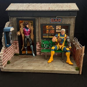 Könnte beinhalten: Ein Diorama mit zwei Actionfiguren vor einer Bar. Eine Figur im Trenchcoat steht im Türrahmen, während eine andere mit einem Metallarm auf einer grünen Bank sitzt. Die Szene umfasst eine Ziegelwand, ein öffentliches Telefon und ein Wellblechdach.