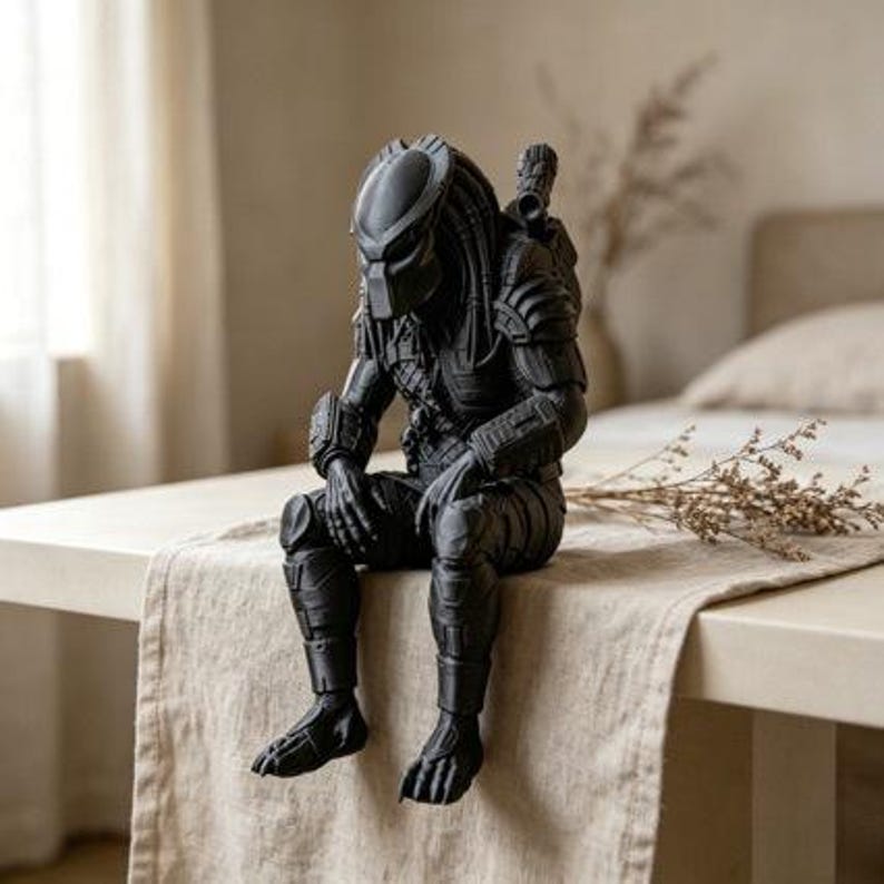 Może przedstawiać: Matowa czarna figurka postaci Predatora z serii film&oacute;w science fiction. Postać przedstawiona jest w pozycji siedzącej z nogami zwisającymi w d&oacute;ł, ubrana w szczeg&oacute;łową zbroję, hełm oraz wyposażona w broń zamontowaną na ramieniu.