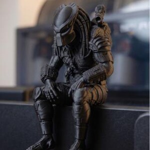 Może przedstawiać: Matowa czarna figurka postaci Predatora z serii film&oacute;w science fiction. Postać przedstawiona jest w pozycji siedzącej z pochyloną głową, z widocznymi detalami pancerza, dredami oraz działkiem zamontowanym na ramieniu.