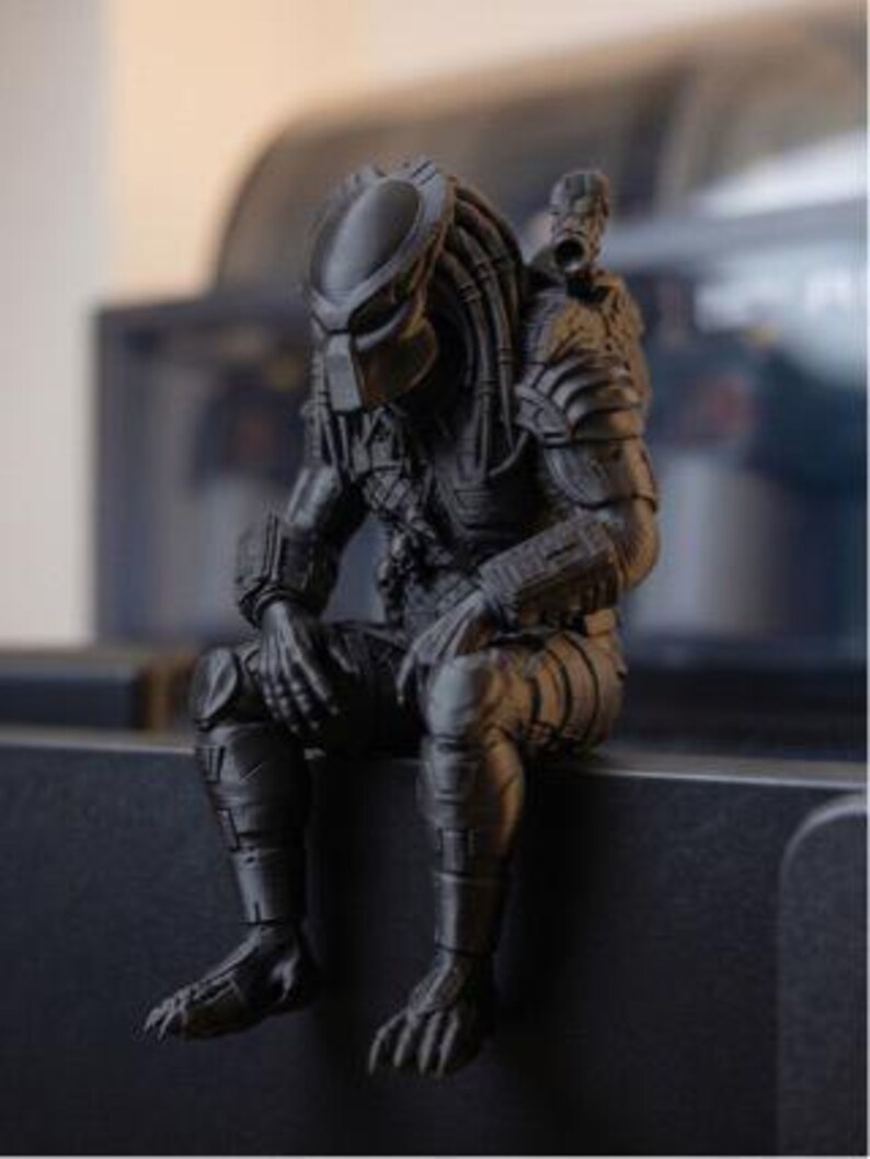 Może przedstawiać: Matowa czarna figurka postaci Predatora z serii film&oacute;w science fiction. Postać przedstawiona jest w pozycji siedzącej z pochyloną głową, z widocznymi detalami pancerza, dredami oraz działkiem zamontowanym na ramieniu.