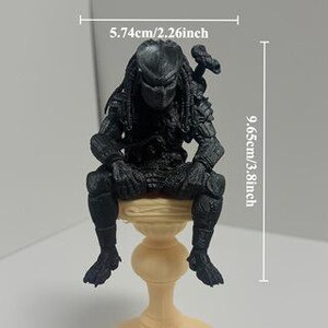 Może przedstawiać: Czarna figurka Predatora siedząca na beżowym postumencie. Postać posiada dredy, zbroję i broń na plecach. Tekst wskazuje wymiary 5,74 cm szerokości i 9,65 cm wysokości, co pokazuje skalę tego przedmiotu.