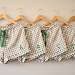 May include: Five pairs of beige and white checkered pajama shorts hanging on wooden hangers. Each pair has a green drawstring and green trim. The shorts are embroidered with the letters A, B, C, D, and E in green.