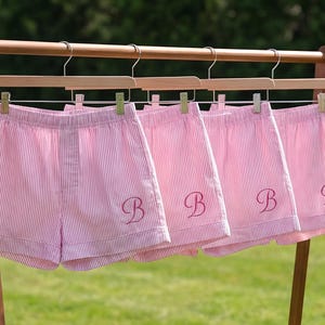 Può includere: Pantaloncini a righe rosa e bianche con la lettera "B" ricamata sul davanti, appesi a un portabiti in legno. I pantaloncini sono esposti su grucce di legno con clip metalliche. Lo sfondo è un prato verde sfocato.
