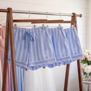 May include: Three pairs of blue and white striped pajama shorts with the letter "J" embroidered on the front. The shorts have a drawstring waist and scalloped hem. They are hanging on a wooden clothes rack.