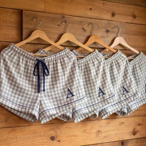 May include: Five pairs of gray and white checkered pajama shorts hanging on wooden hangers. Each pair has a navy blue drawstring and trim. The shorts are embroidered with the letter "A" in navy blue.
