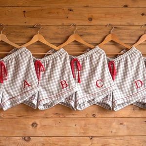 Puede incluir: Cinco pares de pantalones cortos de pijama a cuadros grises y blancos colgando de perchas de madera. Cada par tiene un cordón rojo y una letra bordada en rojo: A, B, C, D y E. Los pantalones cortos se muestran contra una pared de madera.