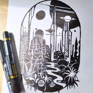 May include: A black and white ink illustration inside an arched frame depicting an astronaut standing in a landscape with alien flora, a winding path, and a planet with rings in the sky. Two black Pentel permanent markers lie to the left.