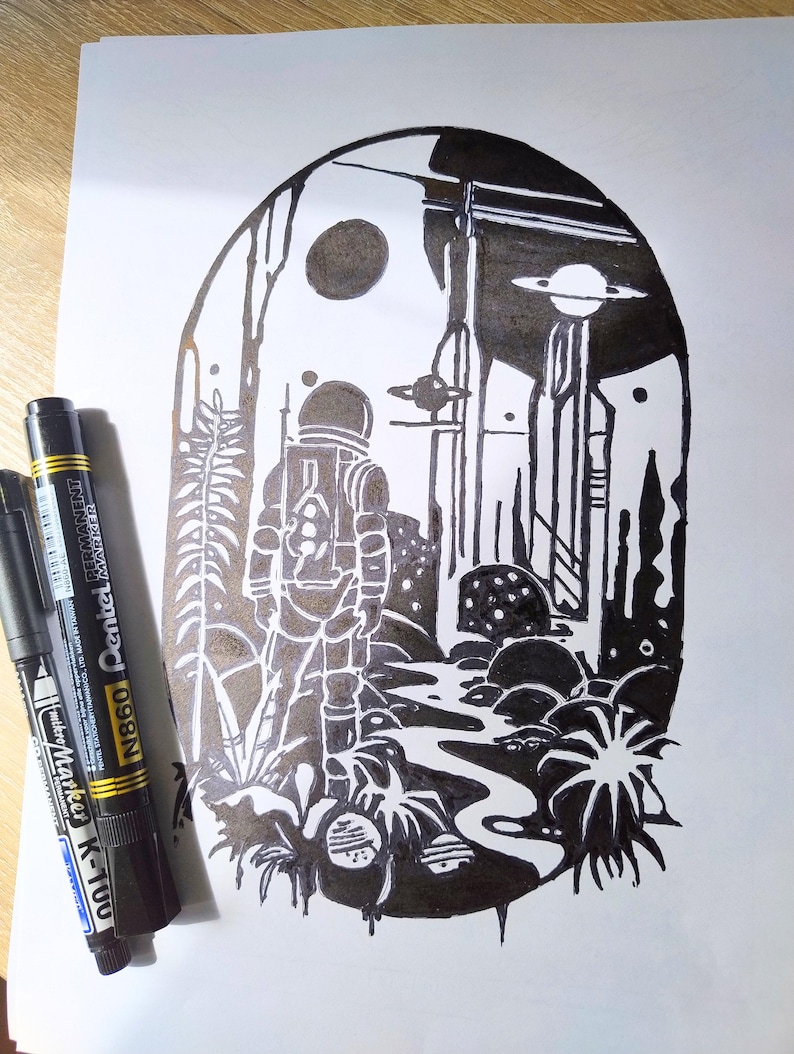 May include: A black and white ink illustration inside an arched frame depicting an astronaut standing in a landscape with alien flora, a winding path, and a planet with rings in the sky. Two black Pentel permanent markers lie to the left.