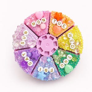 May include: A circular seven-compartment pill organizer with a light purple center. Each wedge-shaped lid is decorated with colorful beads, charms, and white letter beads spelling out the days of the week: MON, TUE, WED, THU, FRI, SAT, and SUN.