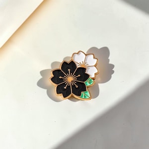Puede incluir: Un broche floral de esmalte con dos flores de cerezo estilizadas. Una flor es negra con centro dorado, la otra blanca. Ambas tienen contornos dorados y están acentuadas con hojas verdes.