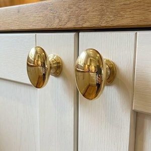 May include: Two polished brass cabinet knobs with an oval, rounded shape. These metallic hardware pieces are mounted onto white, wood-grained cabinet doors, featuring a reflective, smooth surface that catches the light in a kitchen setting.