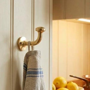 May include: A polished brass wall hook with a rounded end, mounted on a vertical paneled surface. A beige linen-style kitchen towel with blue horizontal stripes hangs from the hook, featuring a fabric loop for suspension on the metal arm.
