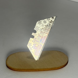 May include: A metallic utility blade featuring an iridescent finish with a repeating pattern of gift boxes and snowflakes, standing upright in a small, oval-shaped wooden base. The blade has a notched edge and a sharp, angled top corner.