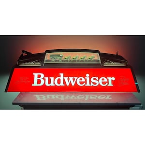 May include: A rectangular Budweiser illuminated sign featuring a bright red background with white lettering. The top section displays a miniature graphic of a horse-drawn wagon. The sign emits a warm glow, highlighting the brand name and logo.