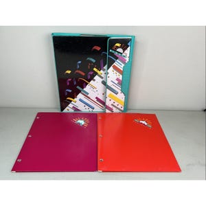 May include: A teal Slapper Binder with a black and white geometric pattern, accompanied by two flat folders in magenta and red. Each item features the Slapper logo, which includes a stylized graphic of a book and the text Slapper.