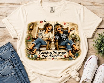 May include: A cream-colored t-shirt featuring a graphic of two people sitting in wooden rocking chairs, holding mugs with text. The design includes the phrase "Rocking Through Life Together" and decorative elements like hearts, flowers, and musical notes.