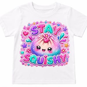 May include: A white short-sleeve t-shirt featuring a colorful graphic of a smiling, glittery dumpling character. The text "STAY SQUISHY" is printed in bold, multi-colored letters above and below the character, surrounded by pink, blue, and purple stars, hearts, and flowers.